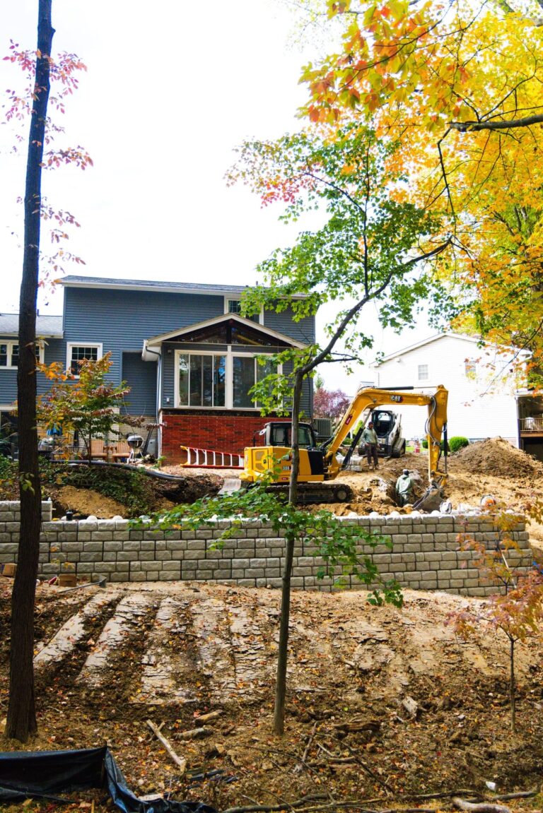 A backyard landscaping project featuring an excavator working near a blue house. The scene includes a partially completed retaining wall, autumn foliage, and visible soil and tree trunks in the foreground. A person stands near the excavator, engaged in the construction work.