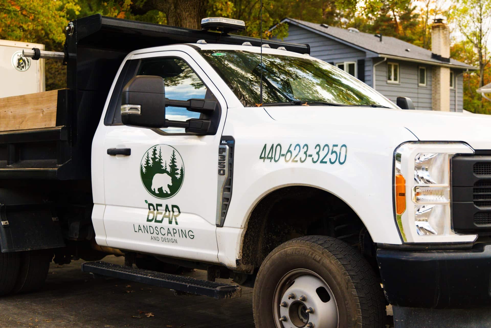 A white landscaping truck with the logo and name 'Bear Landscaping and Design' and contact number on the side, parked in front of a residential house with trees in the background.