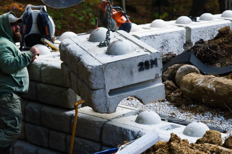 A construction worker in a green hoodie positions a large concrete block using a chain and crane. The block is part of a retaining wall structure, marked with '28M'. Nearby, other construction materials and tools are scattered, including soil and wooden beams.