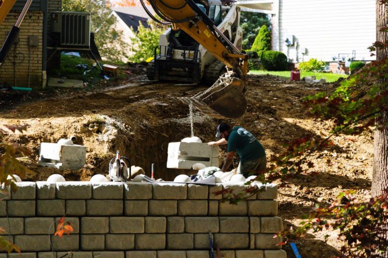 A construction worker in a green shirt supervises a concrete block being lifted by an excavator at a construction site. The site features a partially built retaining wall, and the excavator is positioned on a dirt mound, with trees and a house visible in the background.