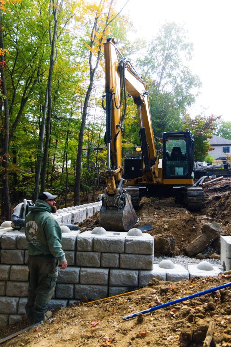A construction scene in a forested area, featuring a yellow excavator working on building a retaining wall made of large, interlocking concrete blocks. A worker in a green hoodie and cap stands nearby, overseeing the project. Trees with green and autumn-colored leaves surround the area, indicating a woodland setting.