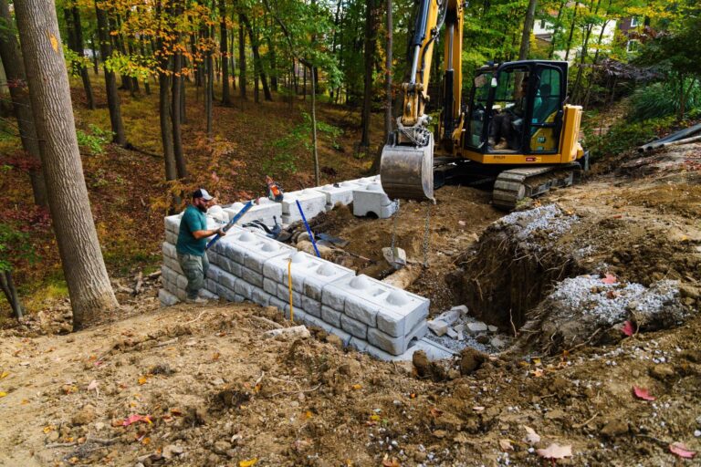 An excavator is positioned on a slope in a forested area, engaged in constructing a retaining wall with large concrete blocks. A worker in a green shirt and cap is using tools to align the blocks. The surrounding area is earthy, with scattered autumn leaves, and tall trees are visible in the background.
