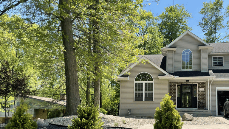 A suburban house with grey siding and large arched windows is partially visible on the right. The entrance features a brick façade and a black front door with glass panels. In the foreground, there is a driveway bordered by small trees and landscaped rocks. Tall green trees with lush foliage are visible throughout the scene, suggesting a serene and wooded neighborhood setting.