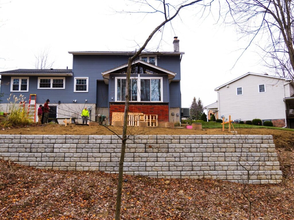 A suburban two-story house with blue-grey siding under construction in the backyard. Several workers are present, with one on a ladder and others arranging materials near the building. A retaining wall made of textured stone blocks is visible in the foreground, with a slope covered in fallen leaves leading up to the construction area. The yard has some mature trees and neighboring houses are visible on either side.