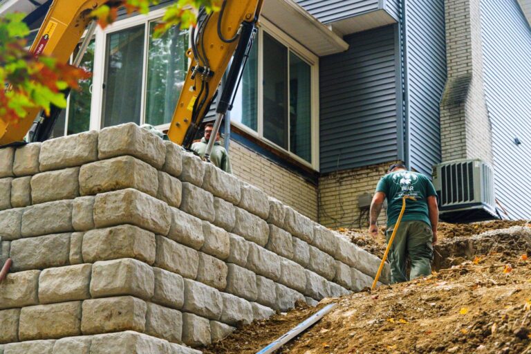 A construction site featuring an excavator near a house with workers building a large block retaining wall. One worker, wearing a green T-shirt, is navigating the soil slope beside the wall, while another operates machinery near a window of the house. The house's exterior includes brick and siding.