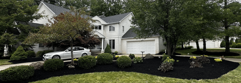 A suburban house with a light gray exterior, a neatly landscaped garden featuring trimmed bushes and a variety of trees. A silver car is parked in the driveway, and the front area is covered with black mulch, creating a well-maintained and organized appearance.