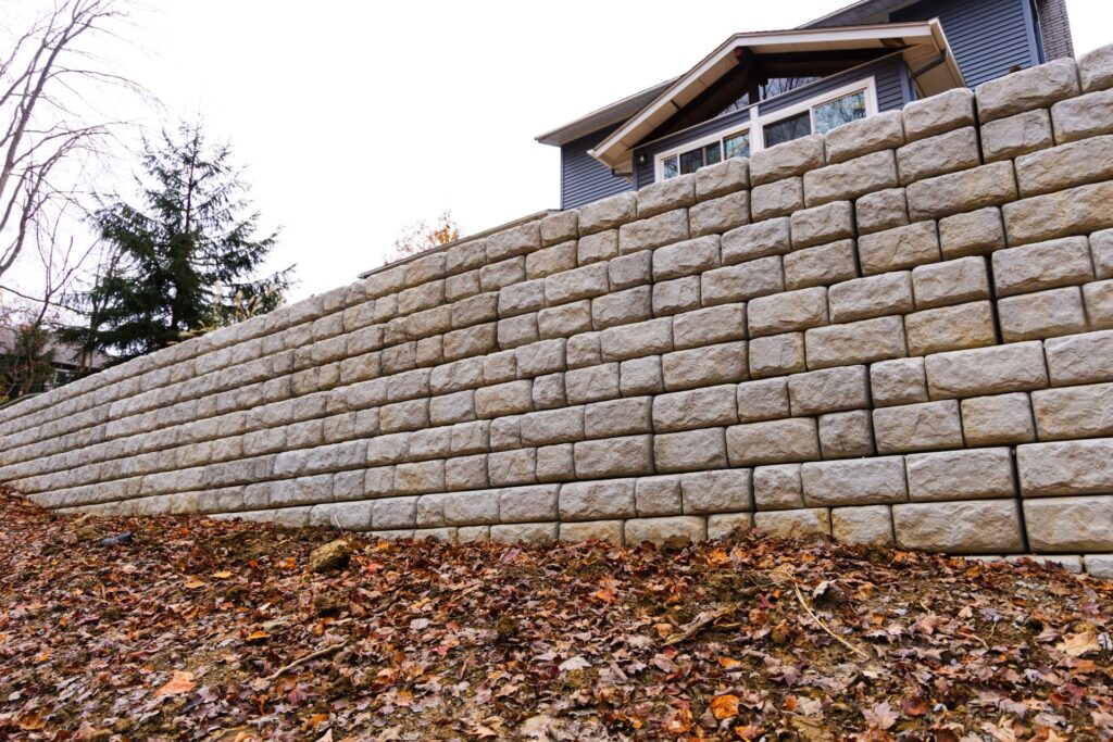 A stone retaining wall with large, rectangular blocks stacked in a grid pattern, extending into the distance. In the foreground, a sloped ground covered with dry autumn leaves. In the background, the upper portion of a house with a blue exterior and a sloped roof is visible, along with the silhouette of a tree.