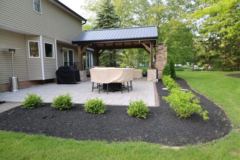 A backyard patio area with a covered pergola attached to a house. The patio is paved with stone tiles and has a table and chairs covered with beige protective covers. The pergola has a sturdy wooden frame with a metal roof. A well-maintained lawn and garden with neatly arranged shrubs surround the patio.