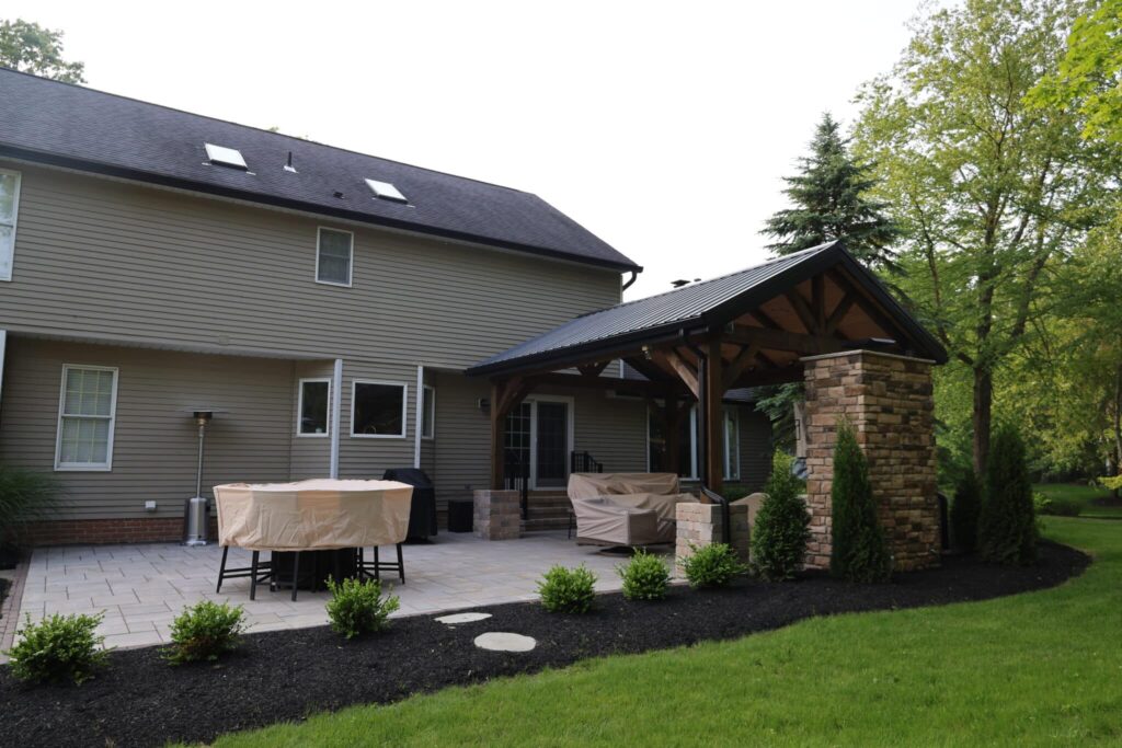 A backyard patio area featuring a covered pergola attached to a two-story house. The patio has stone paving and includes furniture covered with protective covers. Lush greenery and landscaped bushes surround the area, with tall trees in the background.