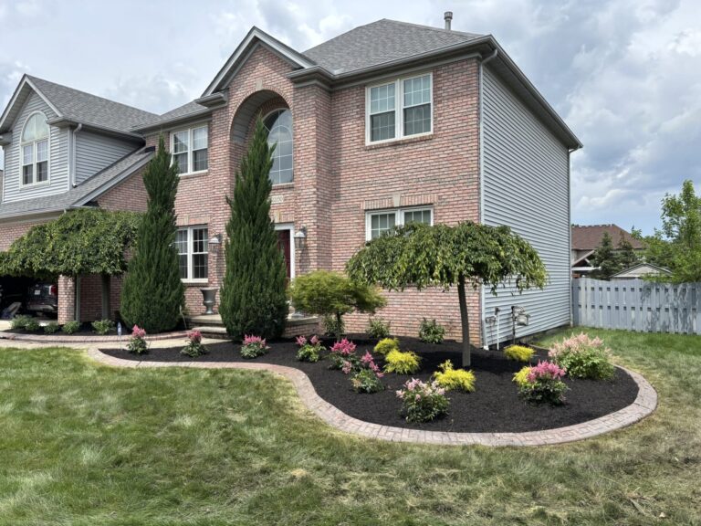 A two-story brick and siding house with a well-maintained front garden. The garden features a variety of plants including tall coniferous trees, colorful flowers, and a neatly trimmed lawn. A brick border outlines a mulched flowerbed with assorted shrubs and ornamental trees.