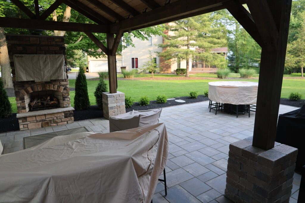 A covered patio area featuring a brick fireplace with a protective cover. The patio has gray stone flooring and is surrounded by a well-maintained garden with green grass and trees. Furniture on the patio is covered with beige protective covers.
