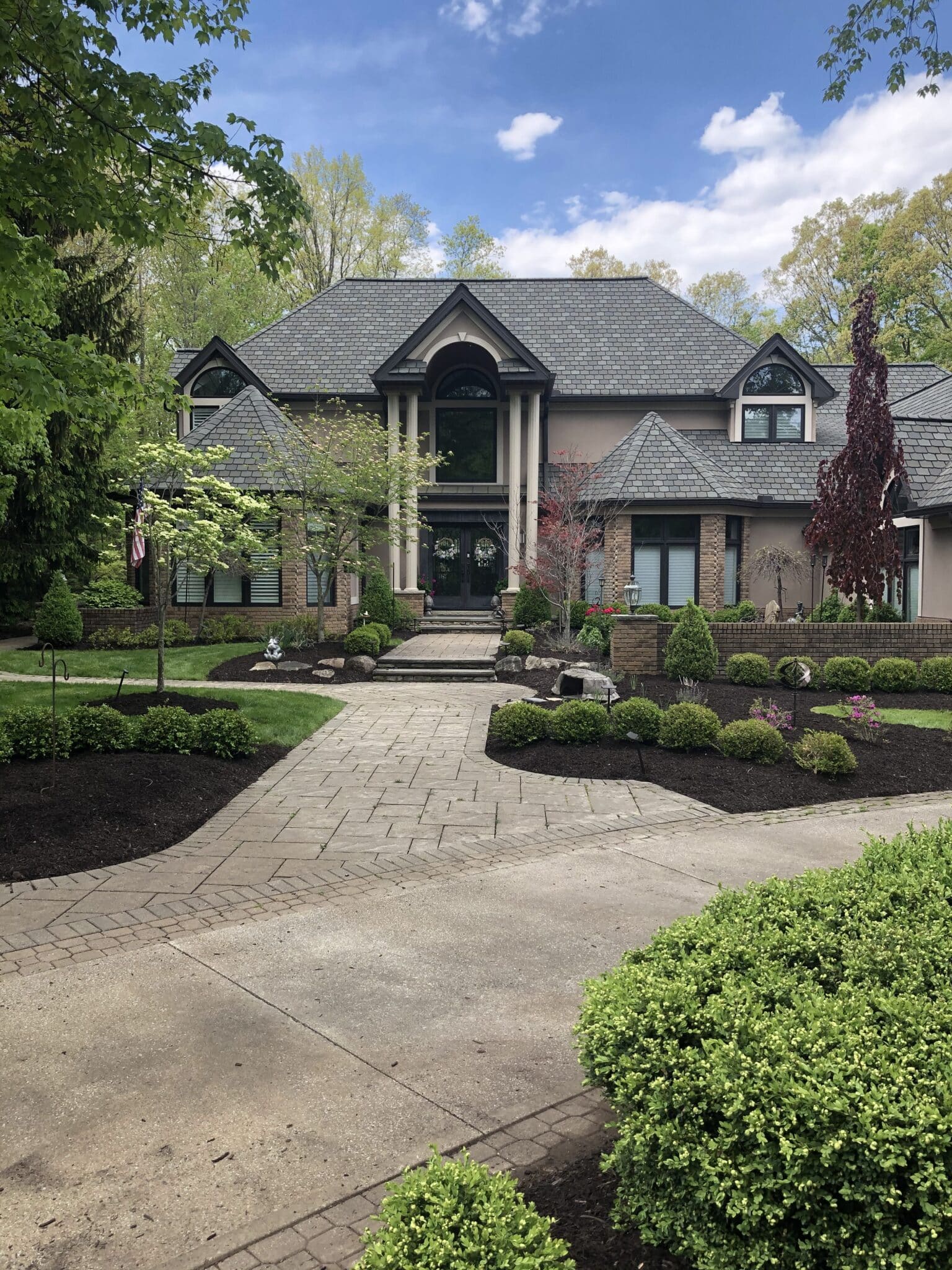 Front view of an elegant, two-story luxury home with a neatly manicured front yard and a paved walkway leading to the entrance. The house features a combination of brick and stucco exterior, large windows, and a sloped roof. The garden is well-maintained with various shrubs, small trees, and mulch beds, all under a clear blue sky.