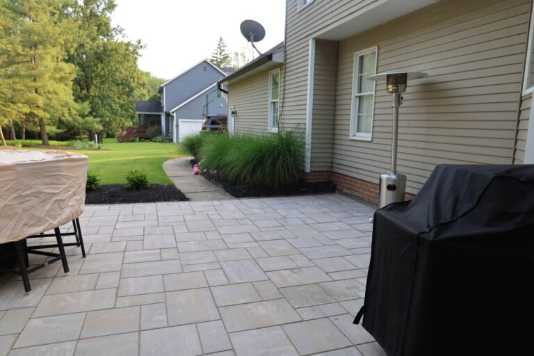 A modern backyard patio with light-colored tiles, featuring a covered grill and a standing gas heater. The patio is adjacent to a beige house with a garden path leading to a well-manicured lawn. In the background, there are trees and additional houses, including a gray house with a visible garage and a vehicle parked in front.
