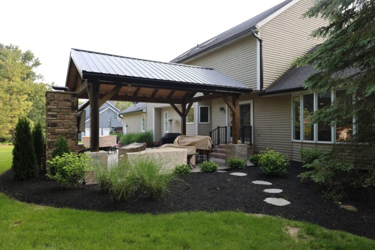 A modern backyard patio with a metal-roofed pergola attached to a beige house with stone accents. The patio features covered outdoor furniture and is surrounded by landscaped greenery and stepping stones leading to a lush lawn.