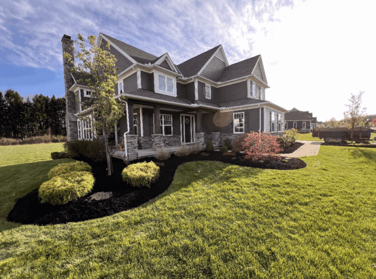 A modern two-story gray house with a sloped roof, featuring a stone chimney and a covered front porch. The house is surrounded by a well-manicured lawn and landscaped garden beds with small bushes and a tree. A paved walkway leads to the entrance, and the scene is bathed in sunlight with a partly cloudy sky overhead.