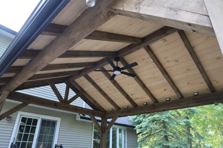 A backyard patio featuring a robust wooden pergola with a natural finish. The pergola has exposed beams and a light-colored wooden ceiling. A modern black ceiling fan is mounted at the center of the structure. In the background, a house with beige siding and sliding glass doors is visible, along with lush green trees.