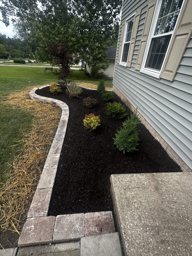 A landscaped garden beside a house with light gray siding, featuring a neatly arranged border of stones. The garden bed is covered with dark mulch and has several small shrubs and plants. A large tree is visible in the background, along with a grassy lawn and a nearby building.
