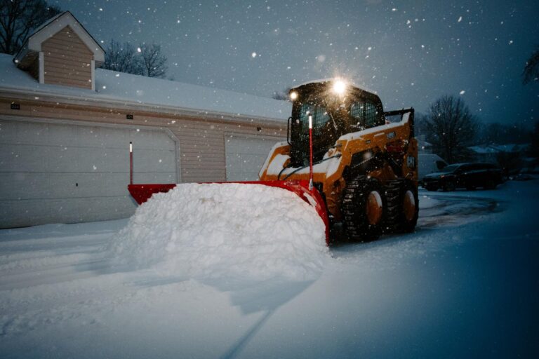 A yellow snow plow with a red blade clearing snow during a snowfall in front of a residential garage with closed doors at night.
