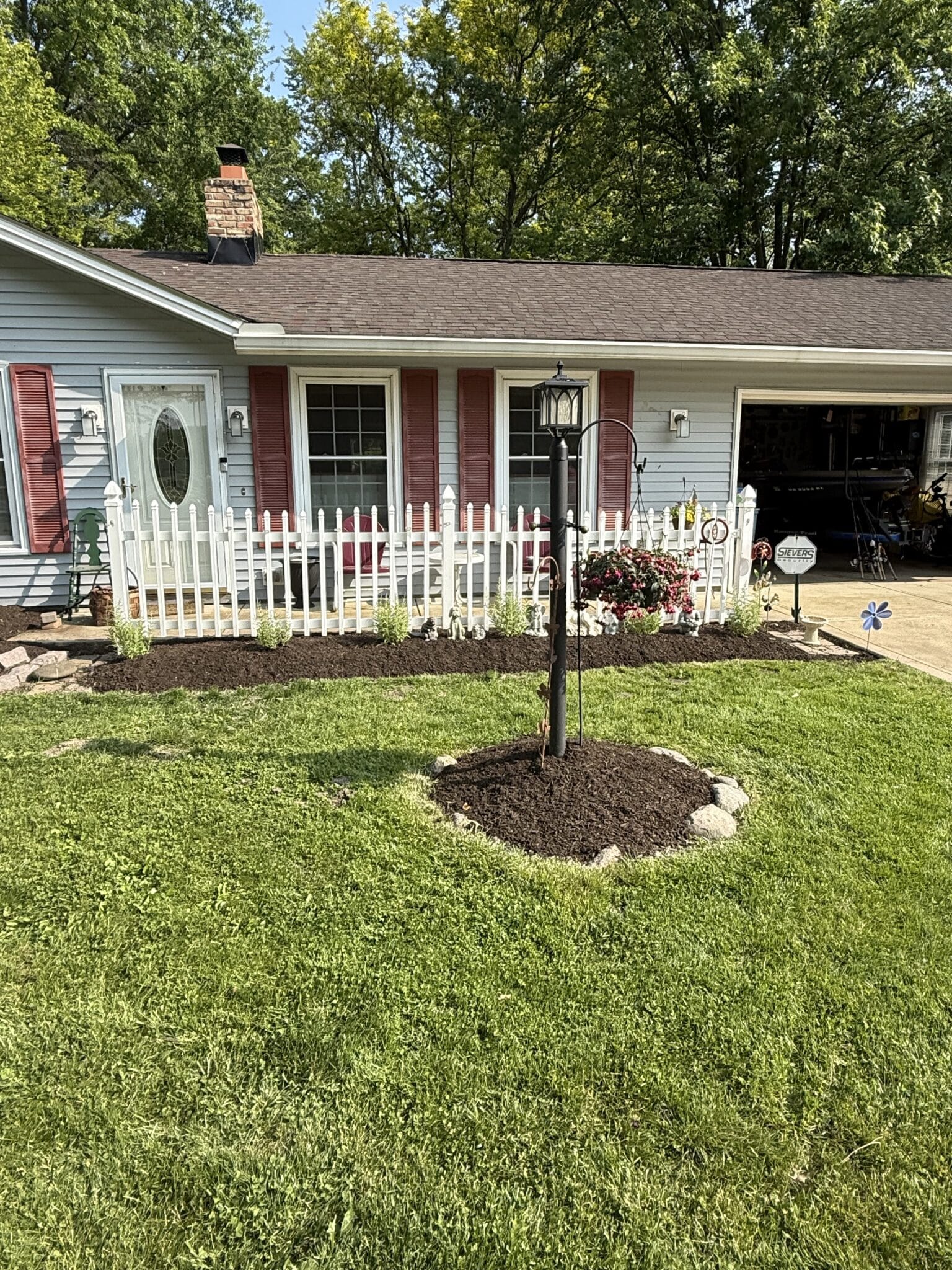 A suburban house featuring light blue siding and red shutters with a small fenced garden in front. A black lamp post is centrally placed on a lawn with fresh mulch. A door with decorative glass is visible, and trees surround the house in the background.