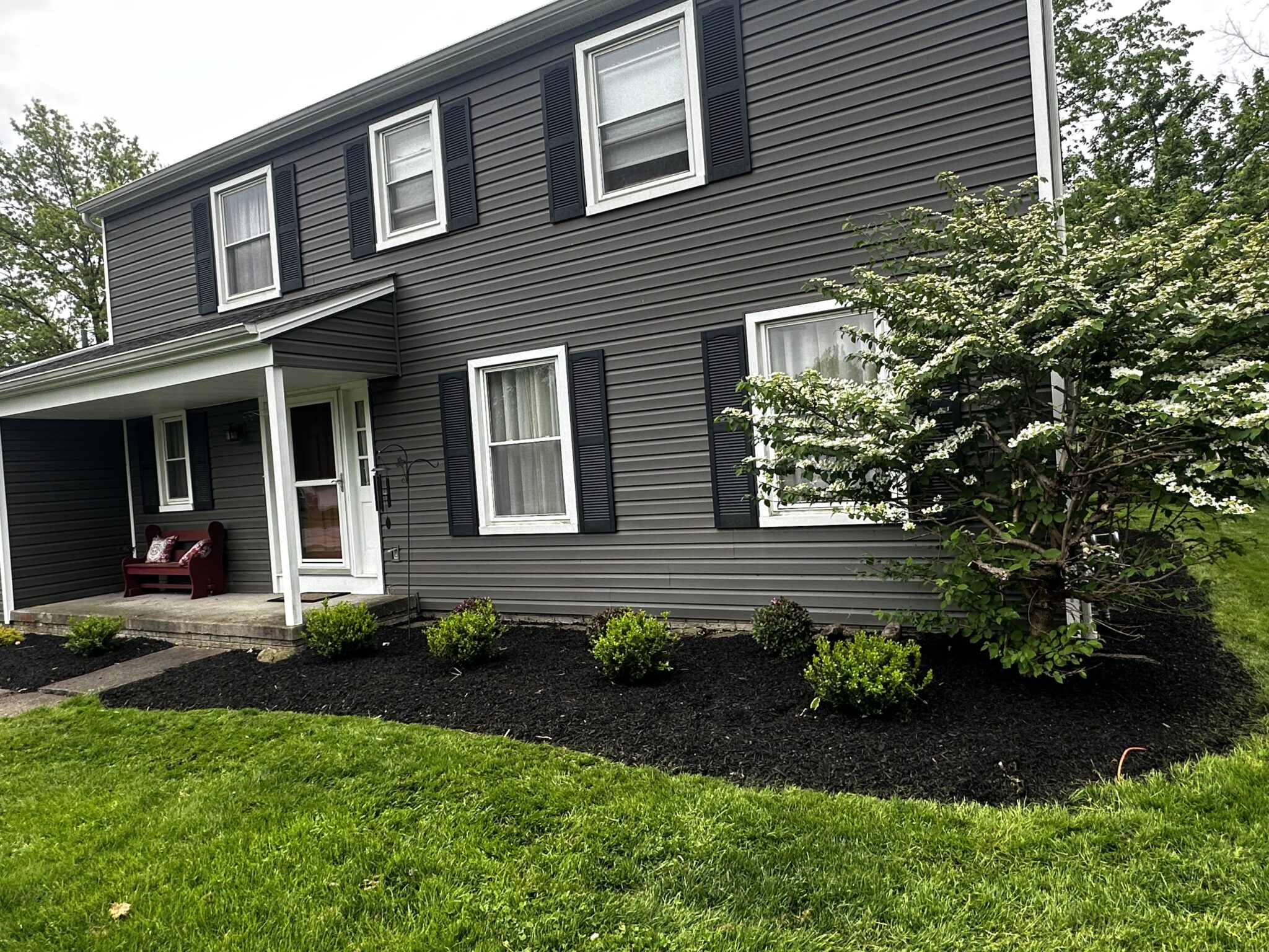 A two-story house with gray siding and black shutters is shown. The house features a porch with a red bench and cushions. In front of the house, there is a garden with small bushes and a flowering tree, surrounded by a grassy lawn.