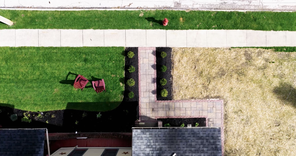 Aerial view of a backyard showing a well-manicured green lawn with two red chairs on the left. A central stone pathway leads to a patio, bordered by small bushes. A sidewalk and fire hydrant are visible at the top of the image, with an area of dry grass on the right.
