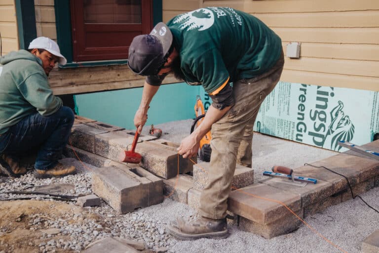 Two construction workers are laying paving stones in front of a building. One worker is kneeling and observing, while the other is using a rubber mallet to position the stones accurately. They are working on a gravel surface and using tools like a level and string line. The background shows a partially finished construction area with insulation and building materials visible.
