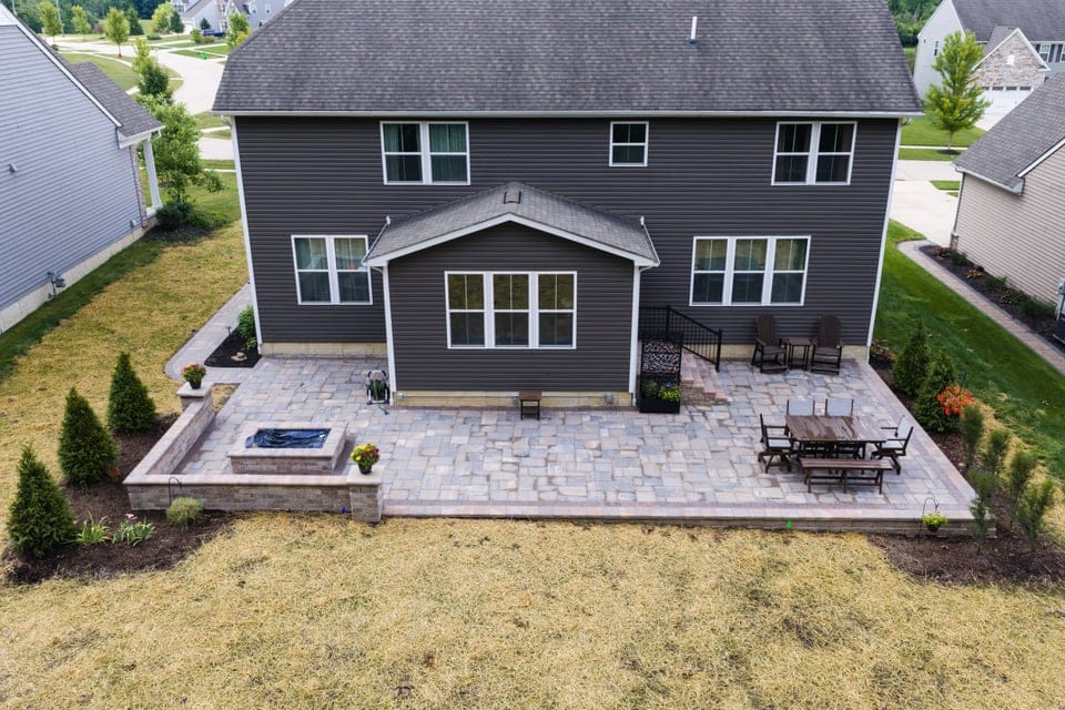Aerial view of a backyard featuring a large patio with stone tiles. The patio includes outdoor furniture, such as a dining table with chairs and a cozy sitting area. There is landscaping with small trees and plants around the patio. The back of a two-story house with multiple windows faces the patio.