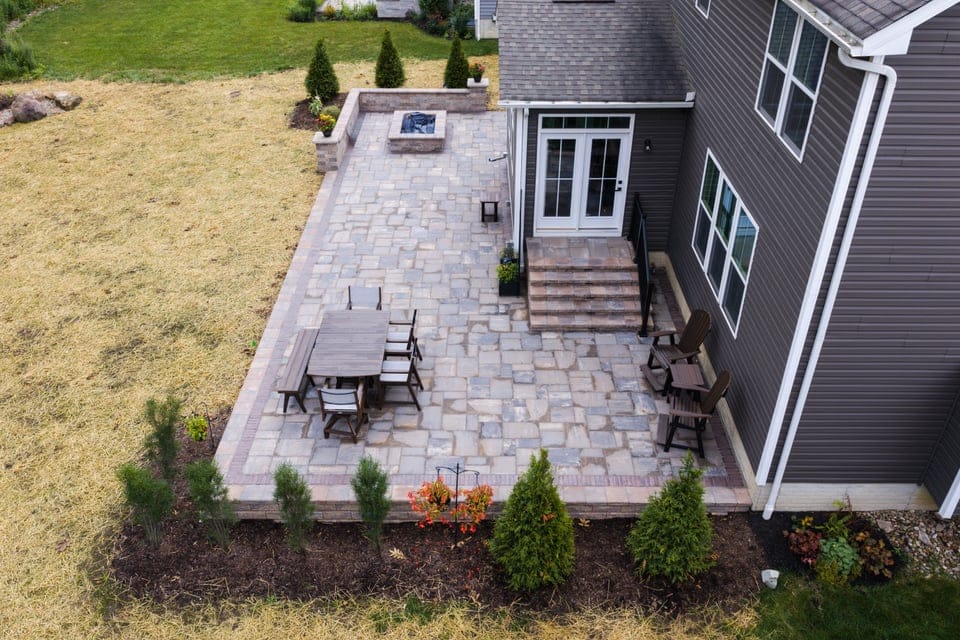 An aerial view of a backyard patio with stone pavers, featuring a wooden dining table with chairs and two Adirondack chairs. A fire pit is located at one end of the patio. The patio is bordered by a garden with small trees and shrubs, and there is a grassy area adjacent to it. The house is visible with large windows and a set of steps leading to the patio.
